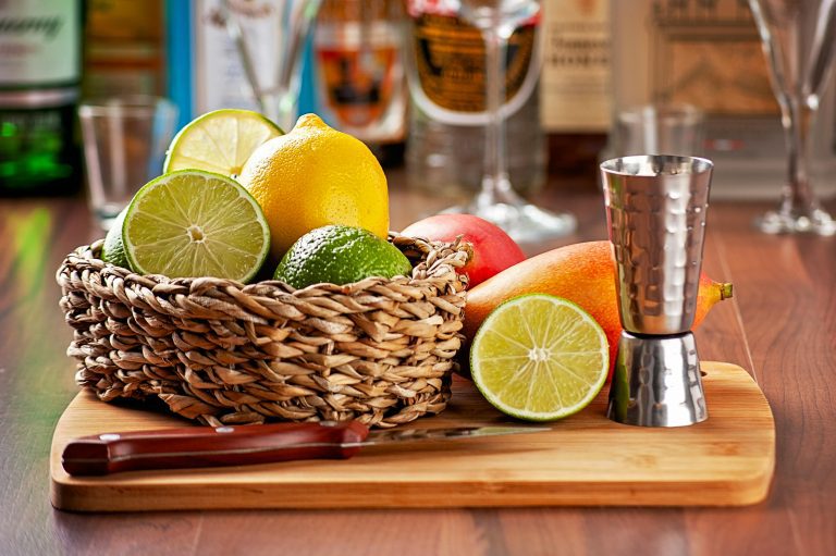 Lemons, limes, and other fruits and vegatables along with a metal cup laid out in a basket and on top of a cutting board