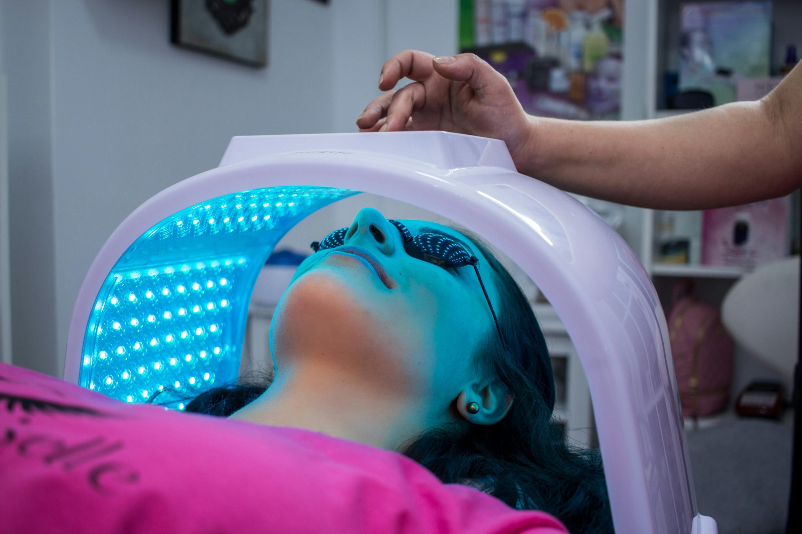 A woman with protective sunglasses under an LED therapy machine filled with glowing blue dot lights. An arm can be seen on top of the machine adjusting the settings.