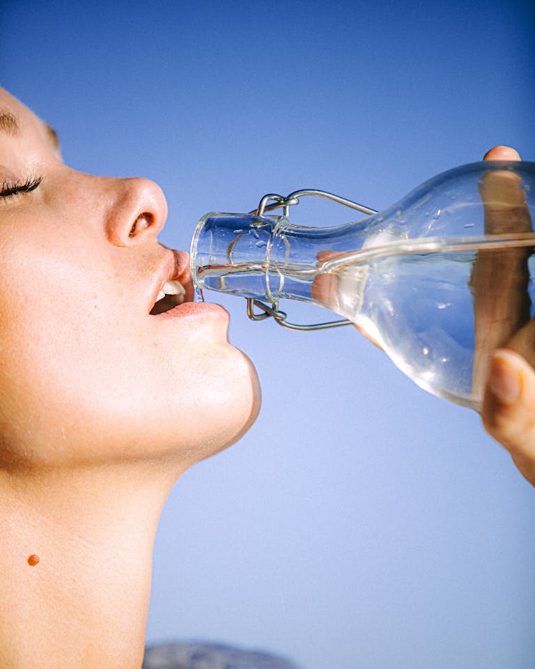 A woman drinking water from a glass bottle against a clear blue sky, emphasizing hydration.